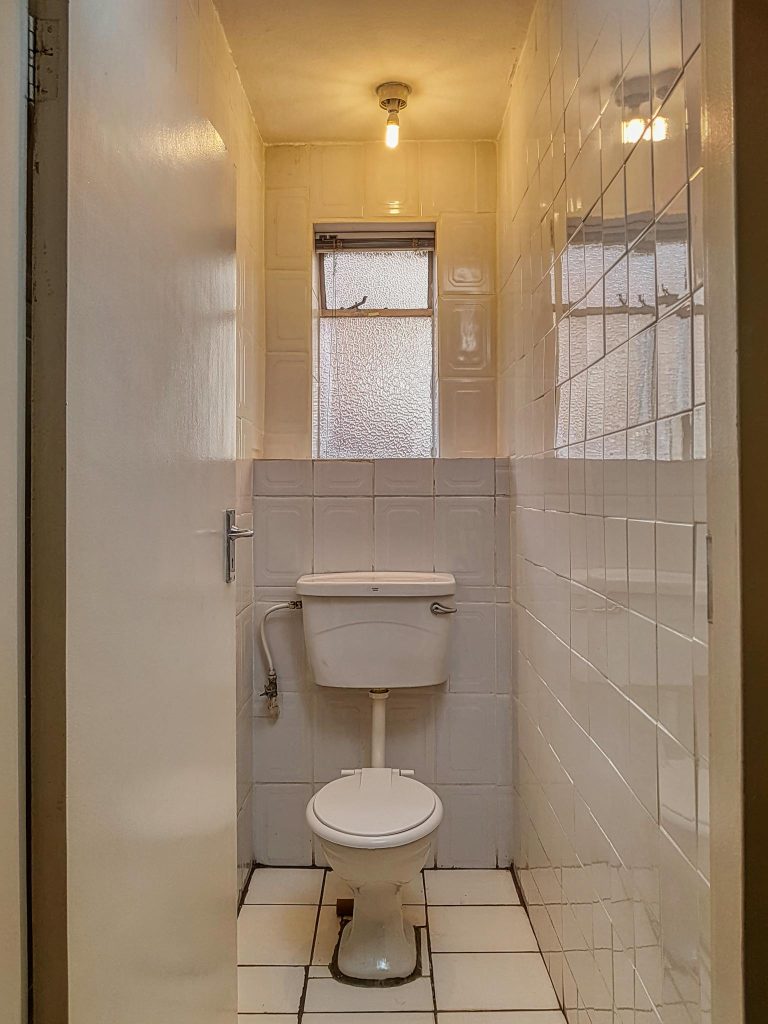 Compact white bathroom with tile walls and vintage toilet, illuminated by warm bulb light.